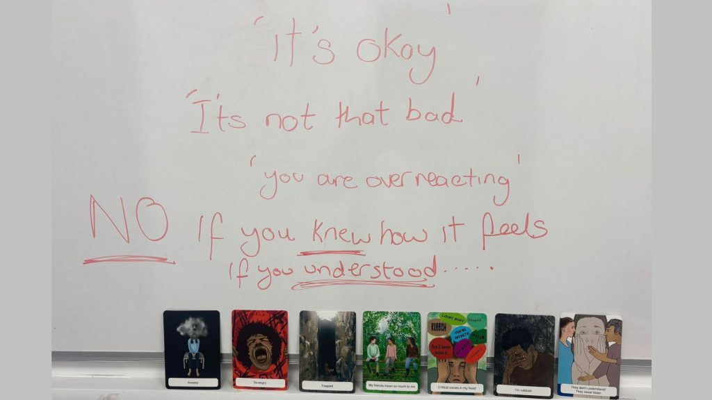 A whiteboard with handwritten red text showing common invalidating phrases. It reads: “‘It’s okay’, ‘It’s not that bad’, ‘You are overreacting.’ NO — If you knew how it feels, if you understood…” with “NO” underlined. Along the bottom of the whiteboard is a row of illustrated emotion cards. The cards depict various feelings, including anxiety, fear, isolation, confusion, and someone shouting, each represented by stylised characters or scenes.